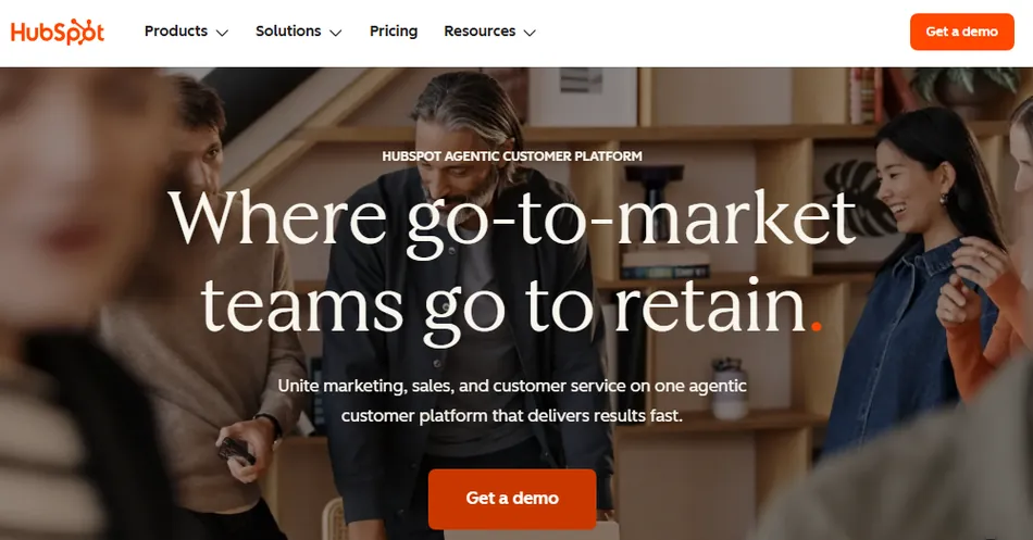 A screenshot of the HubSpot website homepage. The page features a large image of diverse professionals collaborating, overlaid with the text 'Where go-to-market teams go to retain.' Below this, there's a smaller text 'Unite marketing, sales, and customer service on one agentic customer platform that delivers results fast.' The navigation bar at the top includes 'HubSpot' logo, 'Products,' 'Solutions,' 'Pricing,' 'Resources,' and a 'Get a demo' button.